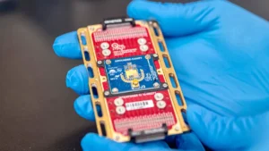 Close-up de um pequeno chip prateado na palma de uma mão enluvada em um laboratório quântico.